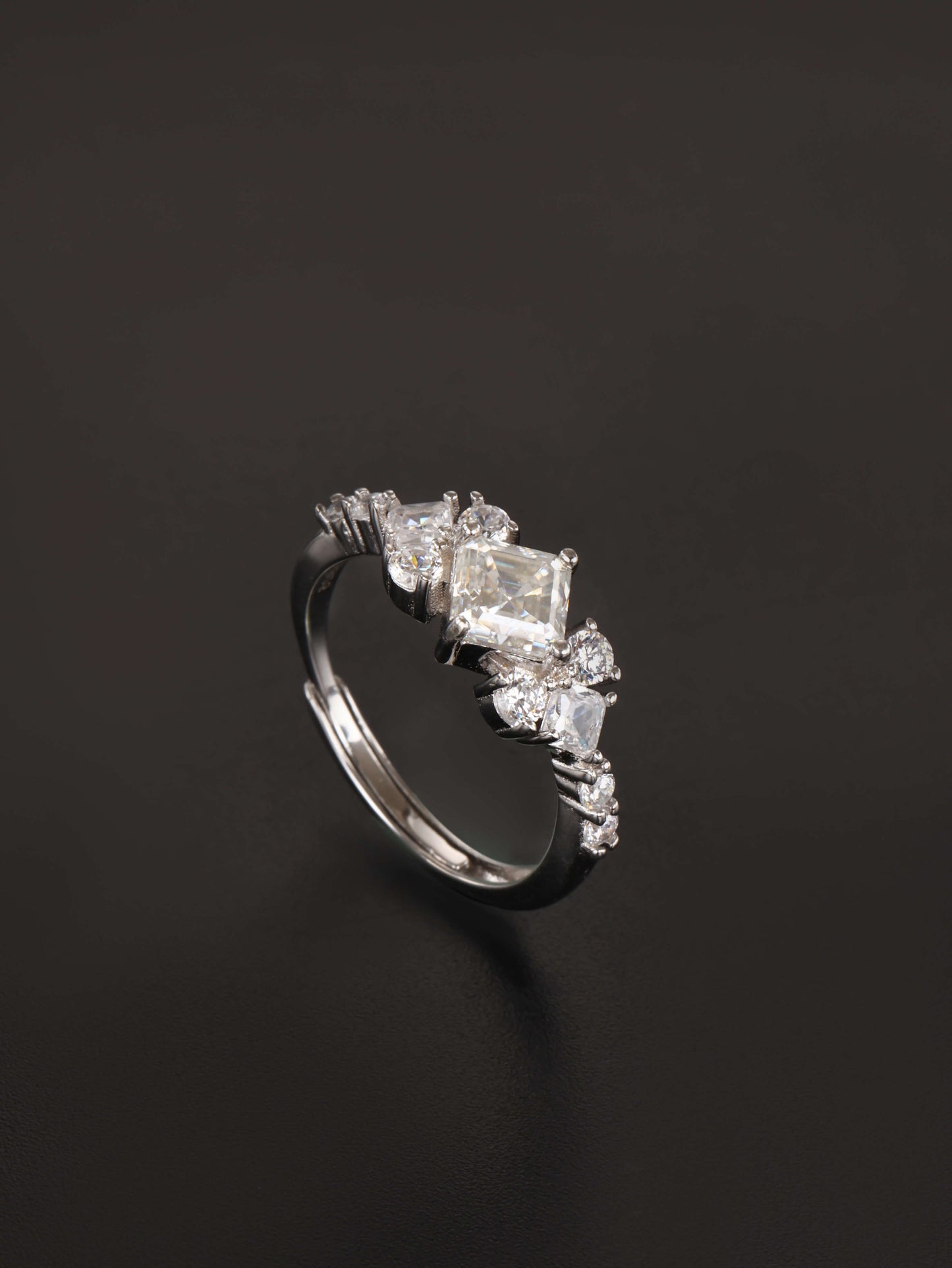 A 925 sterling silver adjustable ring featuring a 1-carat square cut moissanite centre stone in a kite setting, flanked by clustered sparkling side stones, resting on a dark grey background