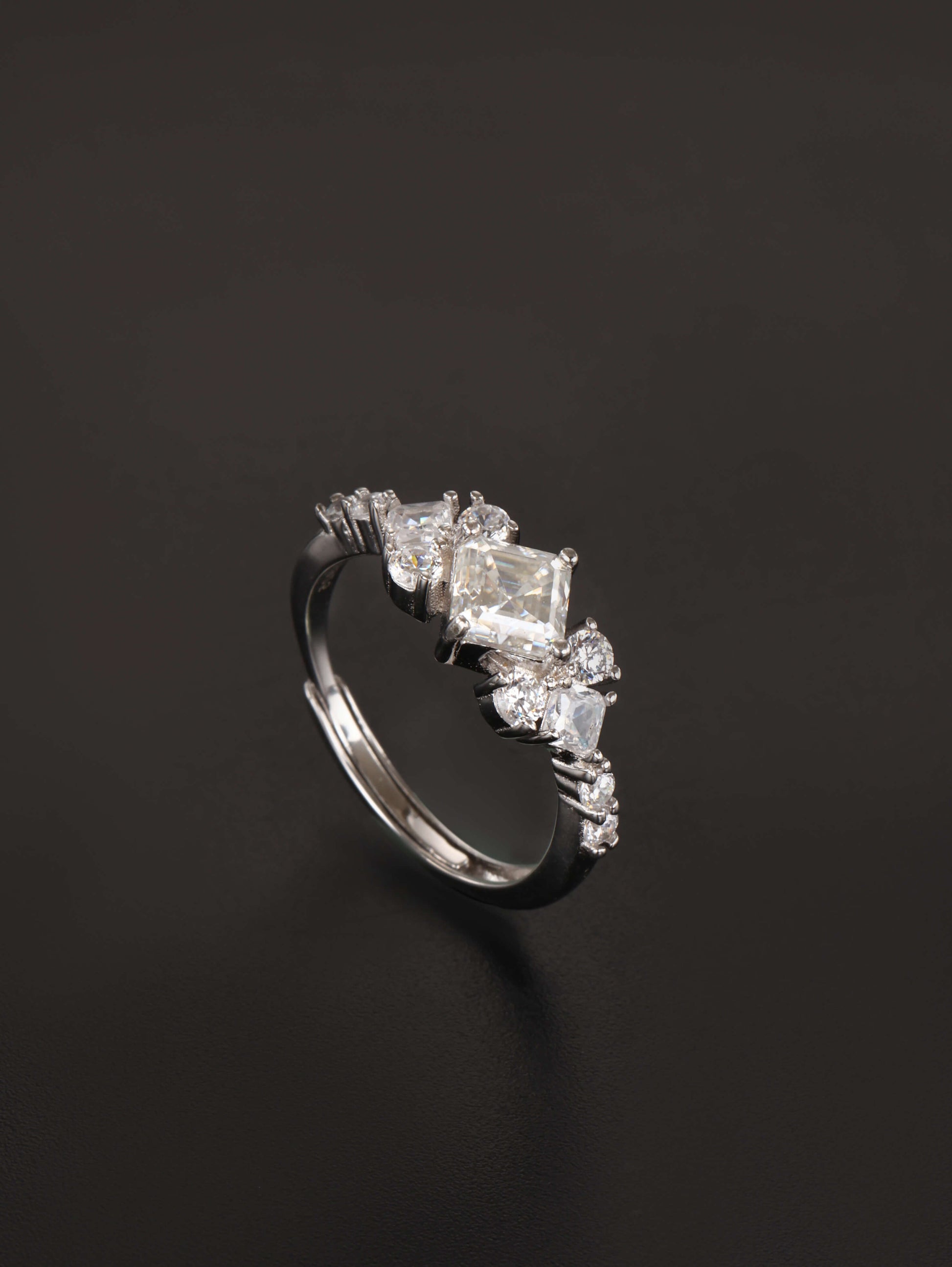 A 925 sterling silver adjustable ring featuring a 1-carat square cut moissanite centre stone in a kite setting, flanked by clustered sparkling side stones, resting on a dark grey background