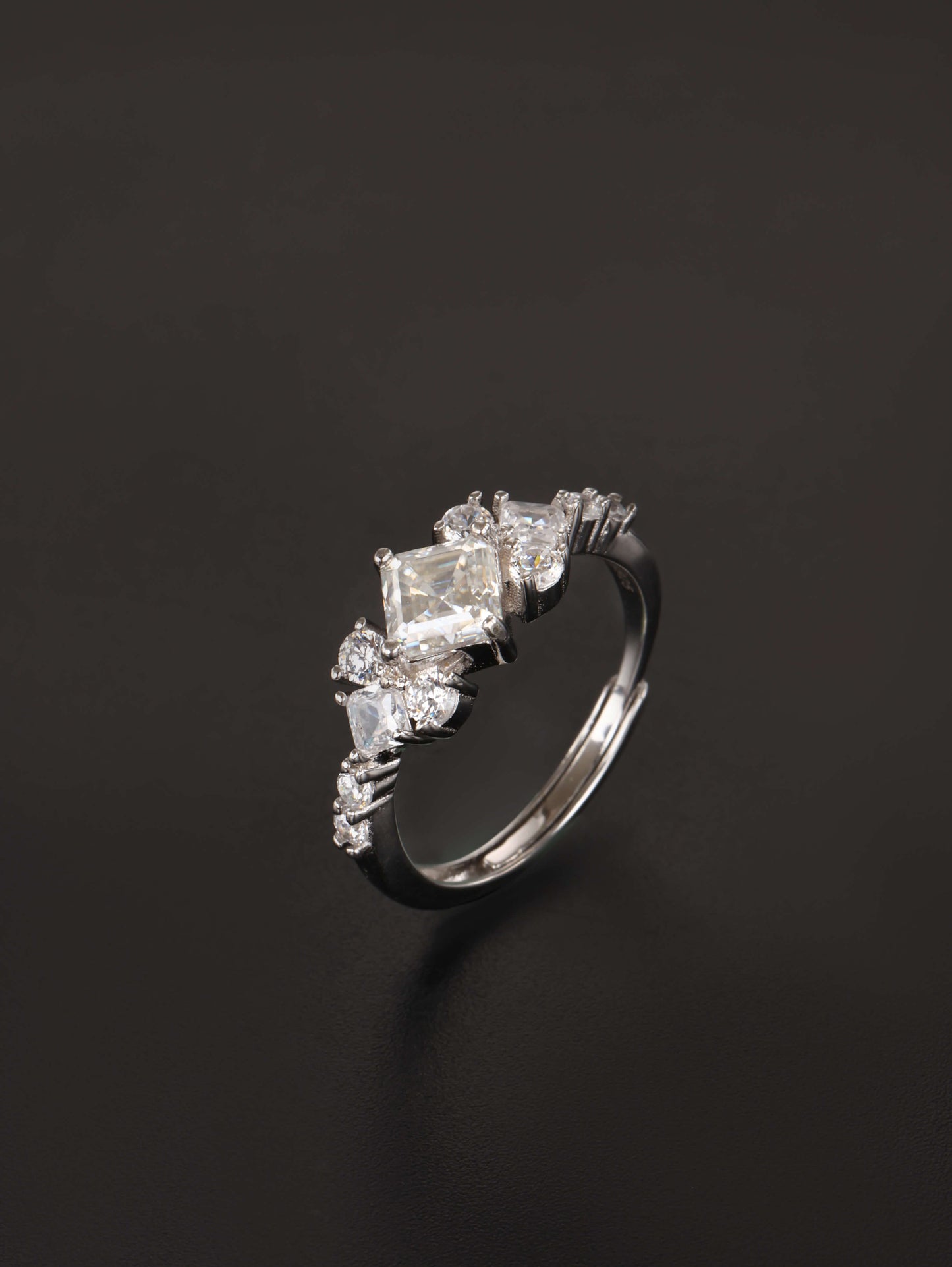 A silver-toned ring featuring a central rectangular clear gemstone surrounded by smaller marquise and round clear stones, photographed on a dark black background.
