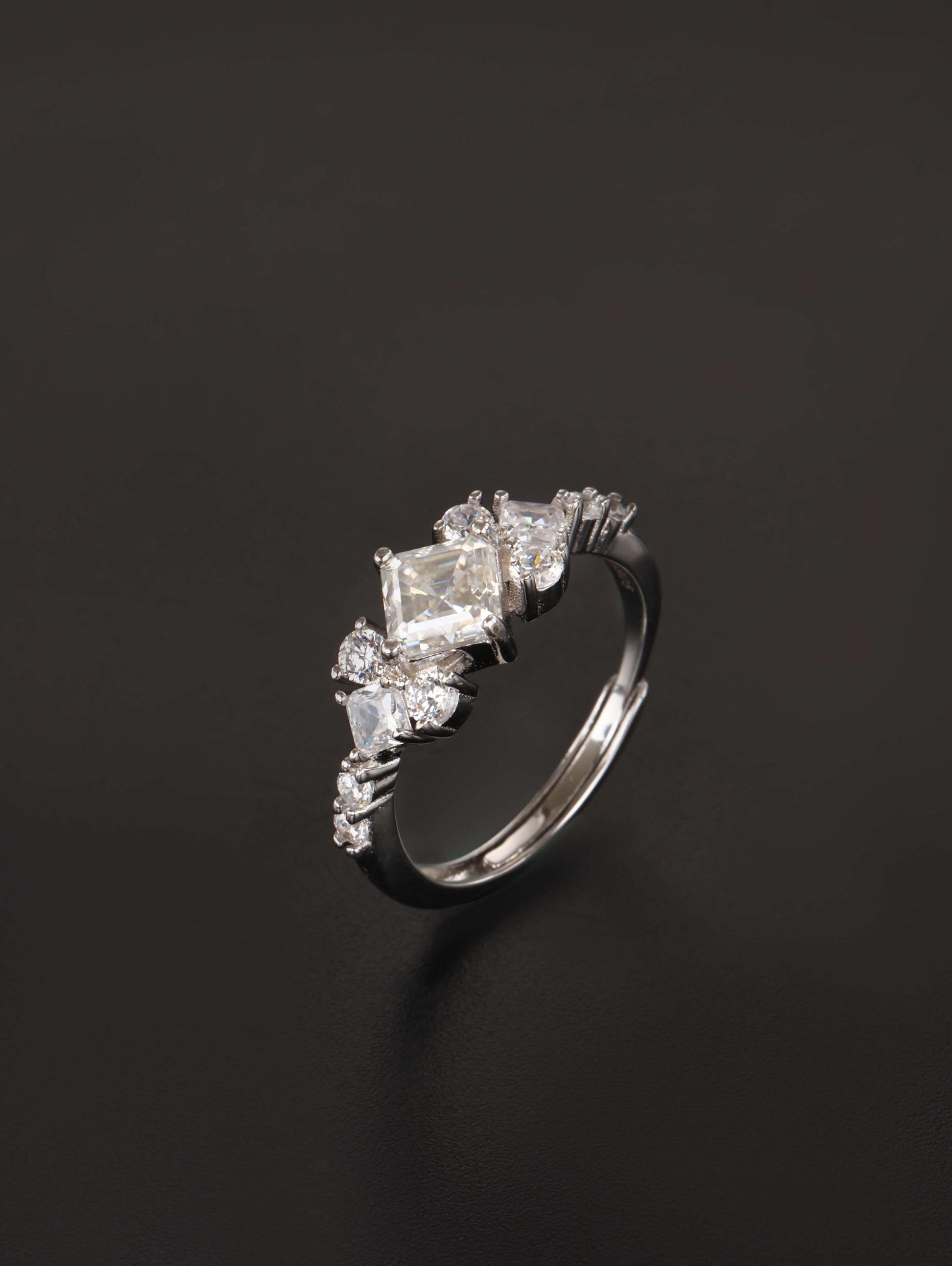 A silver-toned ring featuring a central rectangular clear gemstone surrounded by smaller marquise and round clear stones, photographed on a dark black background.