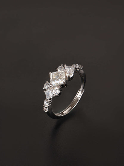 A silver-toned ring featuring a central rectangular clear gemstone surrounded by smaller marquise and round clear stones, photographed on a dark black background.