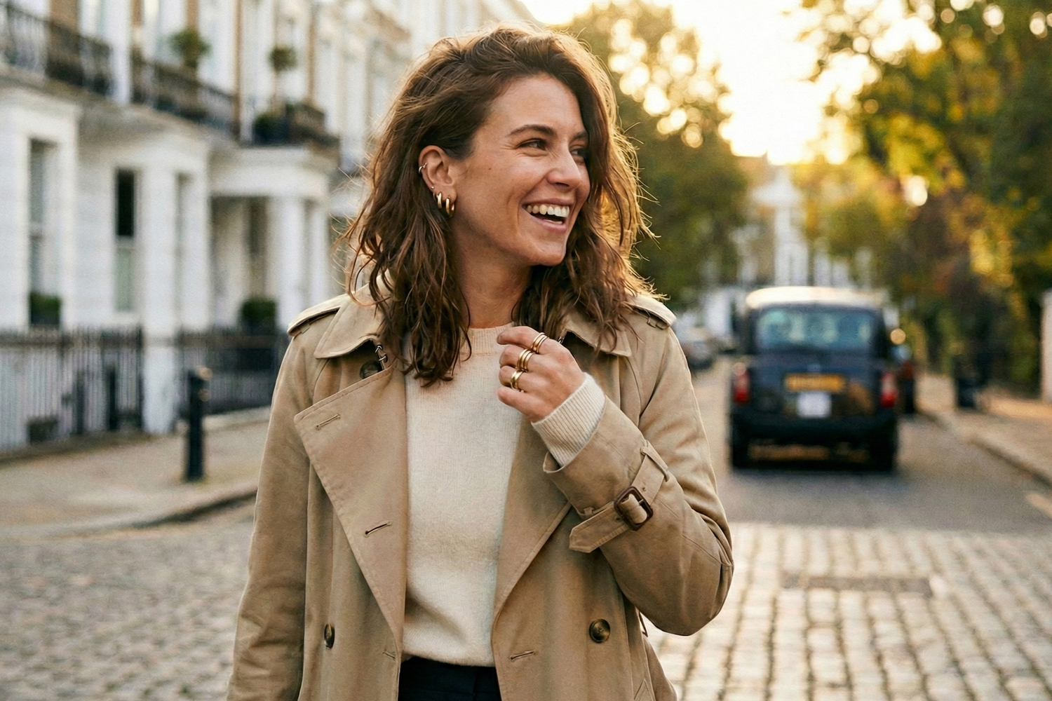 Candid photo of a stylish woman walking in London wearing a beige trench coat. She is modeling Dialuxé 14k gold vermeil ear stacks and stacking rings. Everyday luxury jewellery designed in the UK.